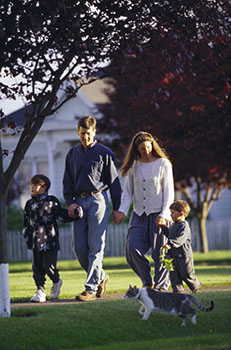 Photograph of a Caucasian family of two young boys holding hands with their mother and father as they walk outdoors with a cat in the foreground